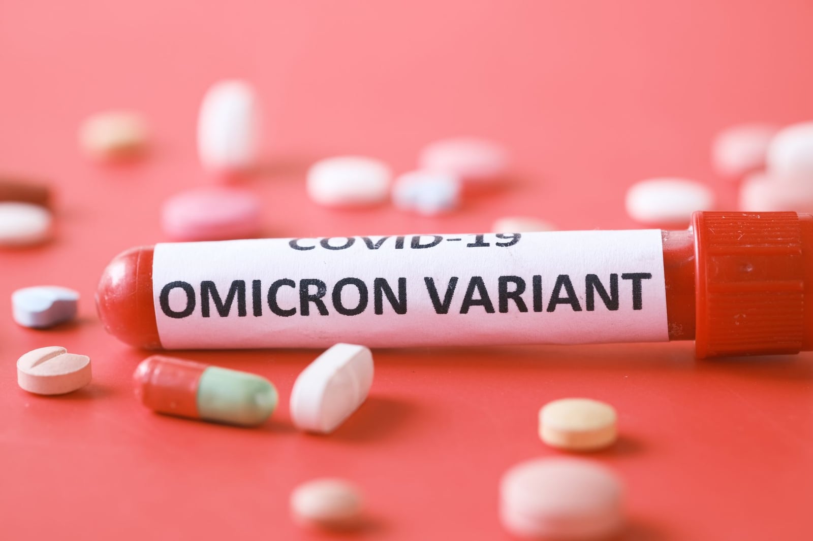 Primer plano de un tubo de ensayo de la variante Ómicron con pastillas variadas sobre un fondo rojo.