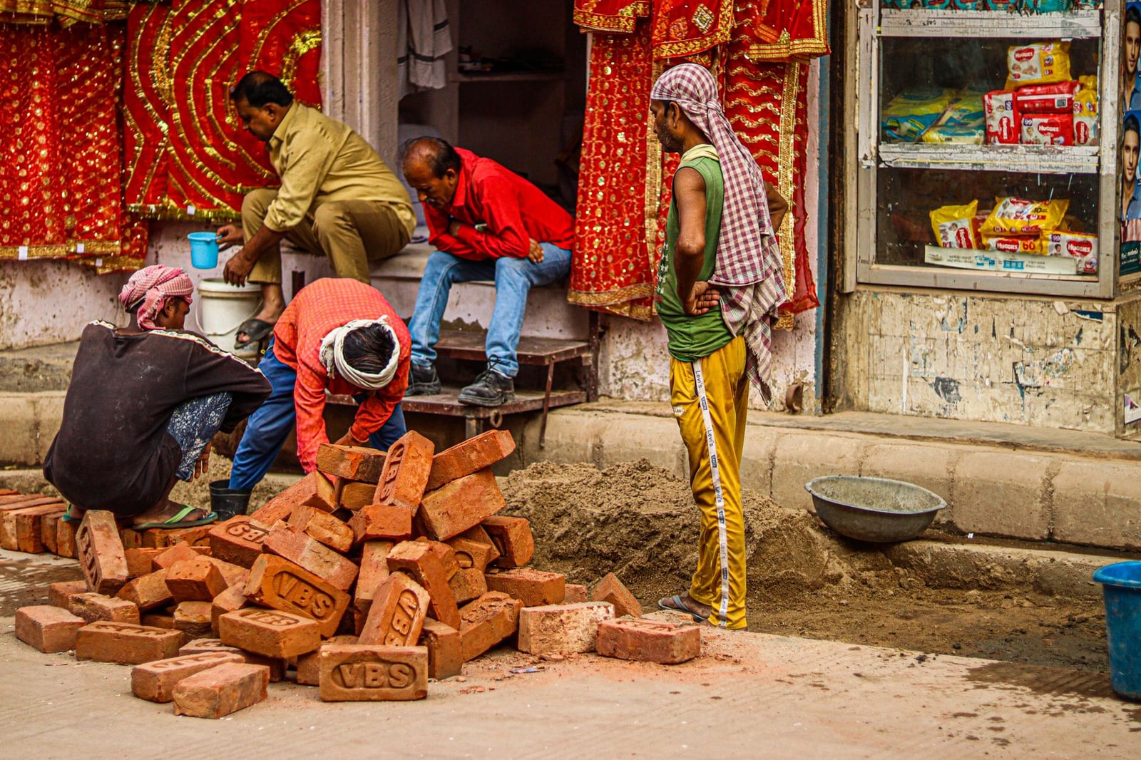 Trabajadores de la construcción manejando ladrillos en una calle bulliciosa, rodeados de textiles coloridos.