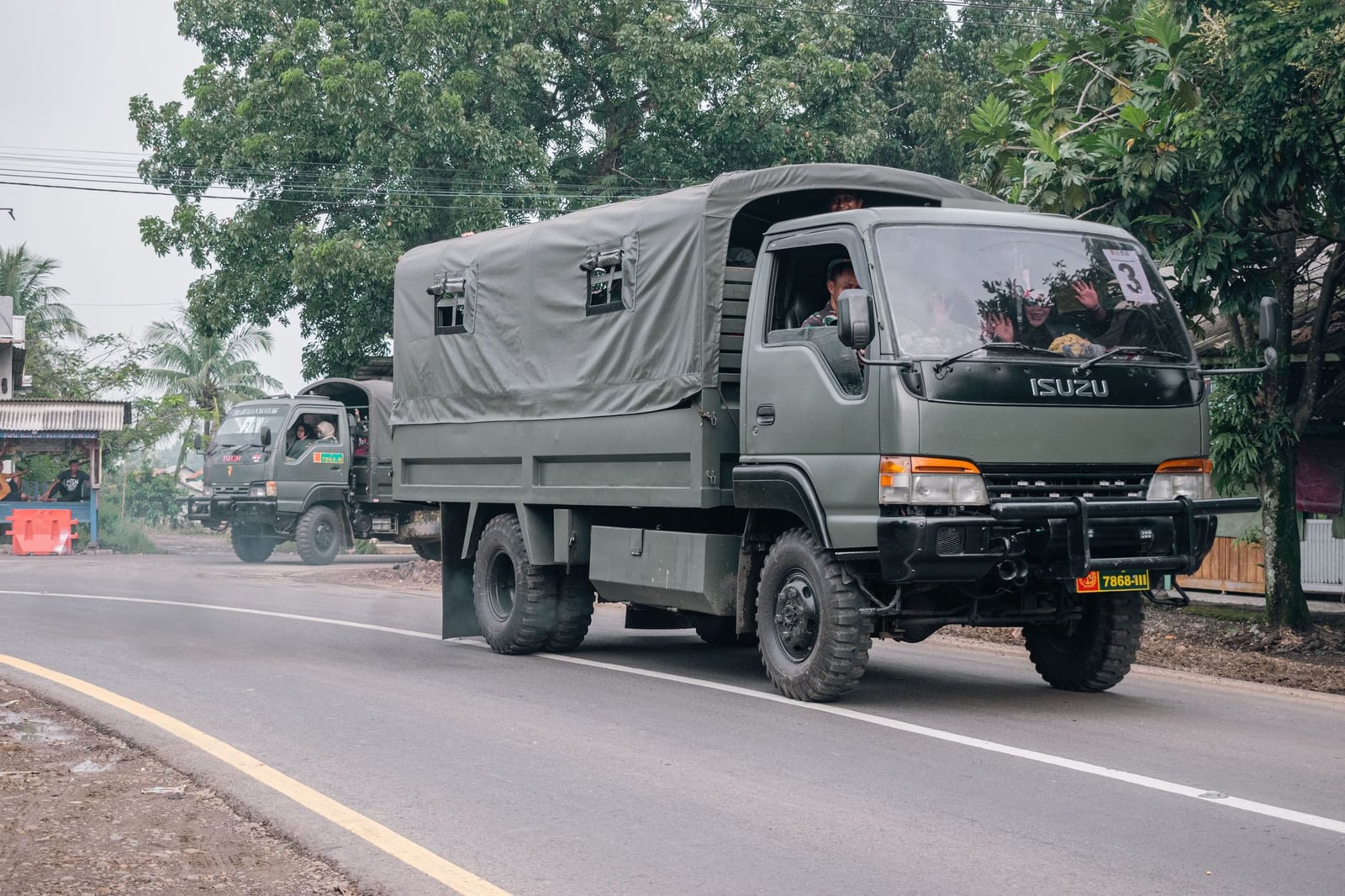 Camiones militares circulando por un camino rural rodeado de árboles y edificios locales.