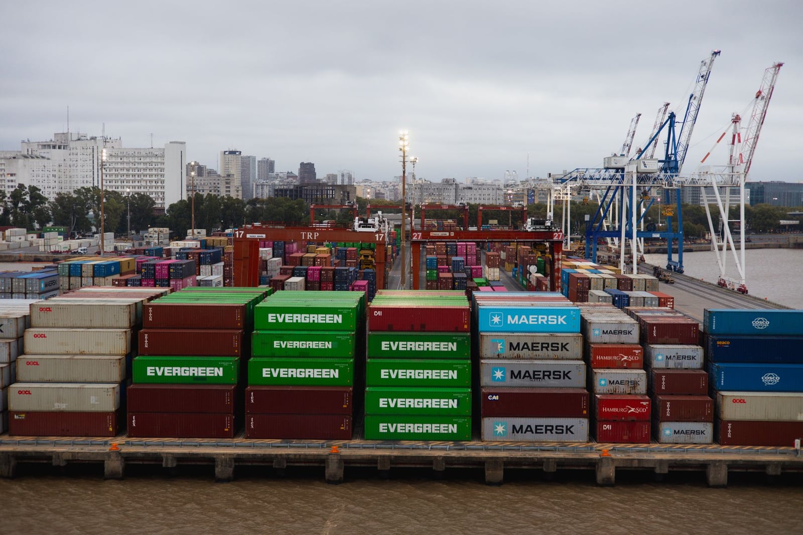 Vista aérea de coloridos contenedores de transporte en un puerto bullicioso con grúas y el perfil urbano de la ciudad.